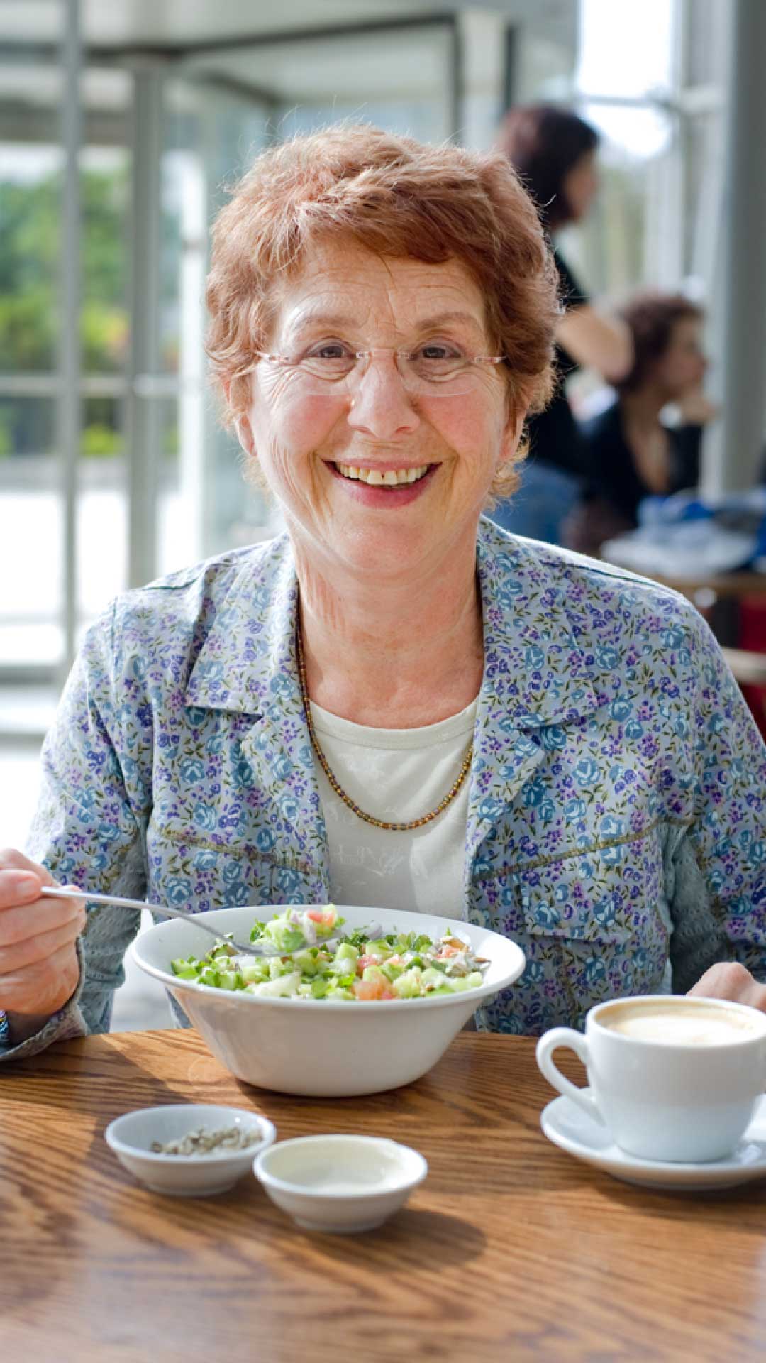 Happy senior smiling while eating a bowl of salad at a table